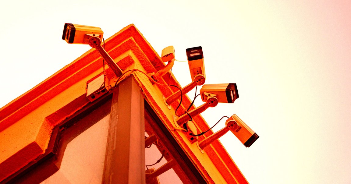 Five security cameras mounted on the corner of a building with an orange hue, angled outward for surveillance. The building has textured walls and a window below the cameras. The sky in the background is clear and light.