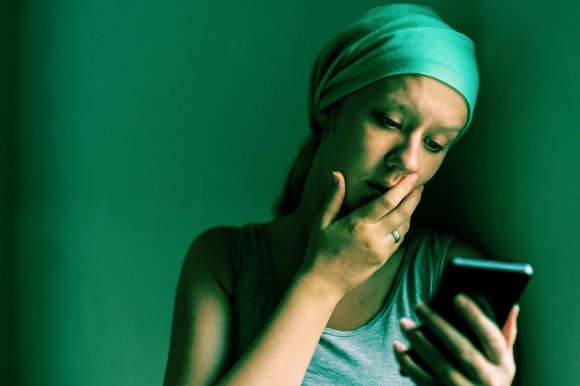 A woman wearing a headscarf and a sleeveless top is looking at her smartphone with a concerned expression, her hand partially covering her mouth. The lighting casts a greenish tint over the scene, emphasizing her contemplative mood.