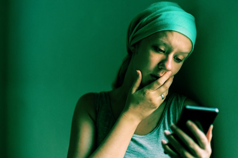 A woman wearing a headscarf and a sleeveless top is looking at her smartphone with a concerned expression, her hand partially covering her mouth. The lighting casts a greenish tint over the scene, emphasizing her contemplative mood.
