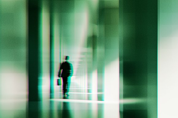 A blurred, abstract image of a person walking down a corridor or hallway with tall vertical columns. The figure appears to be carrying a briefcase and is dressed in dark clothing. The overall color tone is greenish with a glitch or digital distortion effect, creating a futuristic or surreal atmosphere.