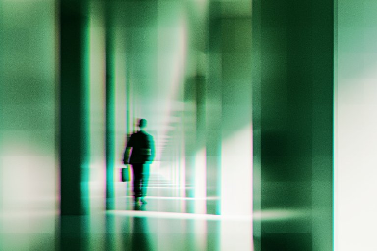 A blurred, abstract image of a person walking down a corridor or hallway with tall vertical columns. The figure appears to be carrying a briefcase and is dressed in dark clothing. The overall color tone is greenish with a glitch or digital distortion effect, creating a futuristic or surreal atmosphere.