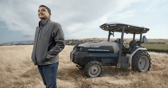 A man wearing a gray jacket and blue jeans stands in a dry grassy field, smiling and looking to the side. Behind him is a modern, compact tractor labeled "MONARCH" with a roof and large tires, set against a backdrop of rolling hills and a partly cloudy sky.
