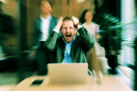 A man in a green suit jacket and blue shirt is sitting at a desk with a laptop, holding his head with both hands and shouting or expressing frustration. Two blurred figures stand behind him in an office setting, with a large plant and window visible in the background. The image has a motion blur effect emphasizing the man's emotional state.