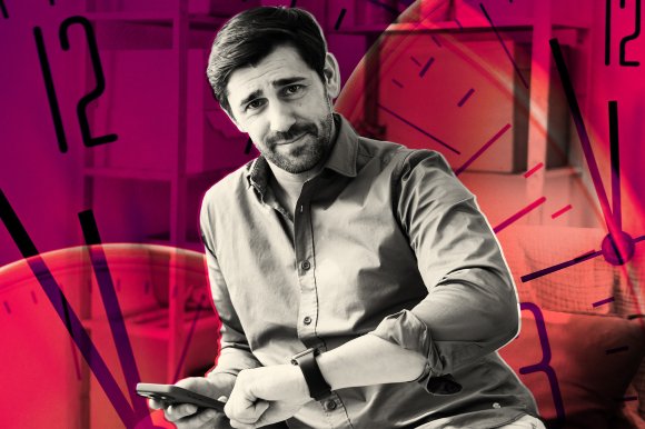 A man with dark hair and a beard, wearing a button-up shirt with rolled-up sleeves, is looking at the camera with a slight smile. He is holding a smartphone in one hand and checking his smartwatch on the other wrist. The background features a large, semi-transparent clock face overlay with bold black numbers and hands, tinted in red and purple hues. The setting appears to be indoors with shelves and furniture faintly visible behind the clock overlay.