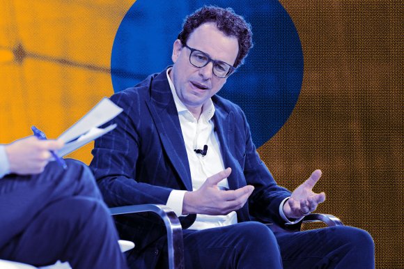 Anthropic CEO Dario Amodei is shown seated, wearing a dark blue blazer over a white shirt. He is gesturing with his hands while speaking. Another person, partially visible, holds a pen and papers on the left side of the image. The background features a large blue circle on an orange and brown textured surface.