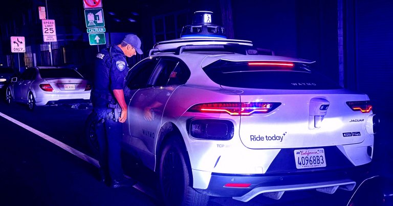 A police officer is standing next to a white Waymo autonomous Jaguar SUV at night, looking inside the driver's side window.
