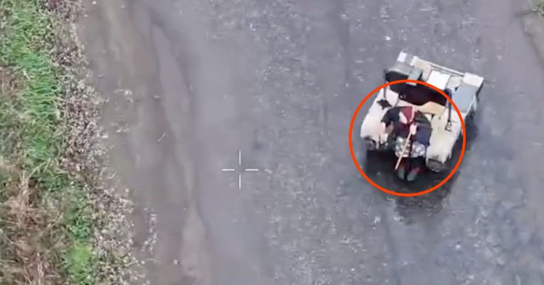 A small, light-colored military robot is shown from above on a paved surface. The robot has an older woman sitting on it.