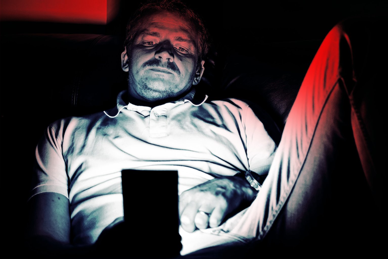 A man wearing a white polo shirt and light-colored pants is sitting in a dark environment, illuminated by the light from a smartphone he is holding. The lighting casts a blueish hue on his face and shirt, with a red light highlighting part of his pants and the background. He has a mustache and short hair, and appears focused on the phone screen.