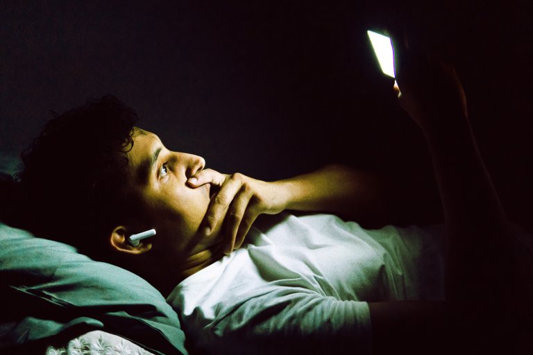 A person lying down in a dark room, illuminated by the light from a smartphone they are holding up. They are wearing a white shirt and wireless earbuds, with one hand resting on their face in a thoughtful pose.