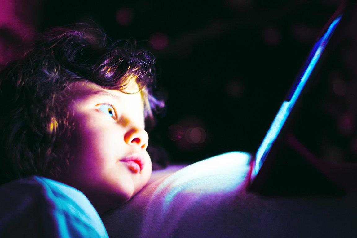 A young child with curly hair is lying down and staring intently at a glowing tablet screen in a dark environment, with the blue light from the screen illuminating their face.