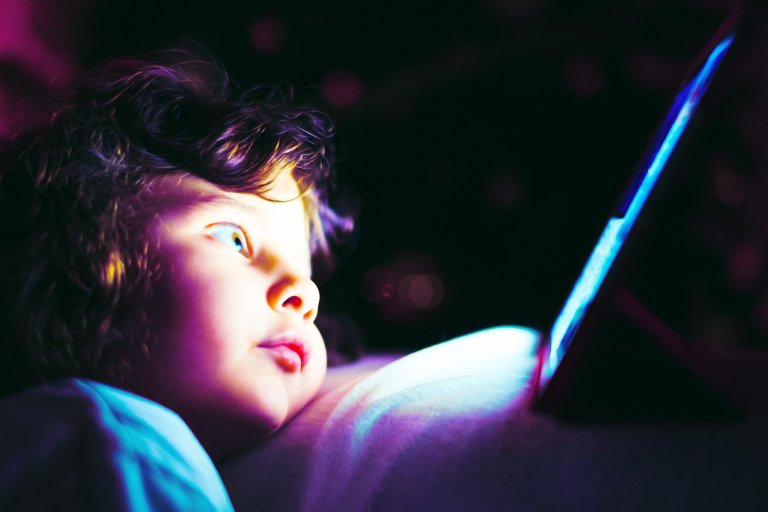 A young child with curly hair is lying down and staring intently at a glowing tablet screen in a dark environment, with the blue light from the screen illuminating their face.