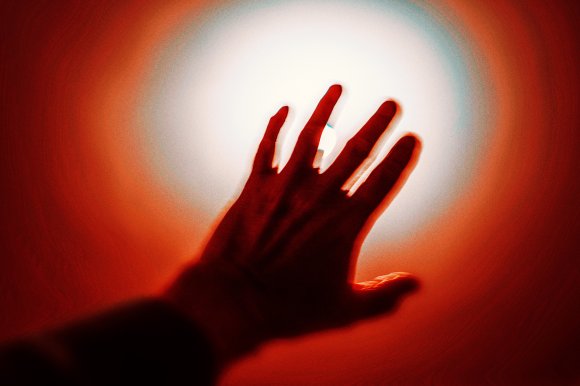 A hand reaching towards a bright white light source, surrounded by a red and orange glowing aura. The image has a dramatic, high-contrast effect with the hand appearing as a dark silhouette against the intense light.