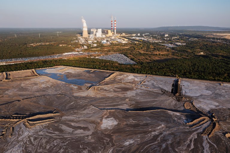 A large industrial power plant with multiple cooling towers and two tall smokestacks emitting white smoke is visible in the distance. In the foreground, there is a vast area of disturbed land with muddy, water-filled pits and earth-moving equipment. Surrounding the disturbed land are dense green forests, and beyond the power plant, there are more industrial buildings and infrastructure. The sky is clear and blue.