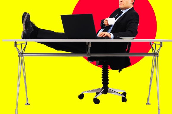 Man in a black suit sitting on an office chair with his feet up on a modern desk, holding a white coffee cup and working on a laptop, against a bright yellow background with a large red circle.