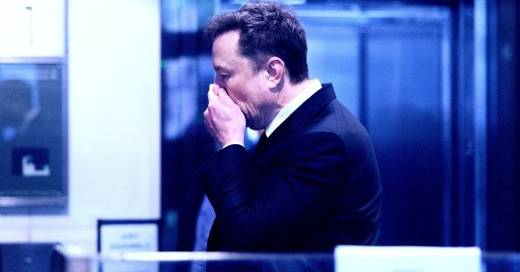 Elon Musk is shown in a dark suit and white shirt, standing indoors, covering his mouth with his hand. The background is blurred with blue tones, suggesting an office or formal setting.