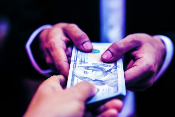 Two hands exchanging a stack of US 100-dollar bills, with one hand giving and the other receiving the money. The background is dark, and the hands are in focus, highlighting the transaction.