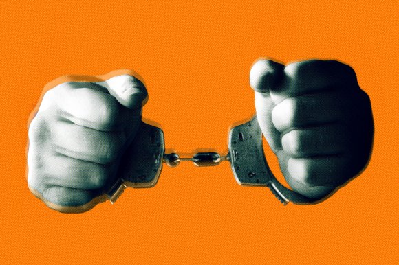 Two clenched fists in black and white are shown wearing handcuffs against a solid bright orange background. The image has a high-contrast, stylized effect with a slight shadow around the fists.