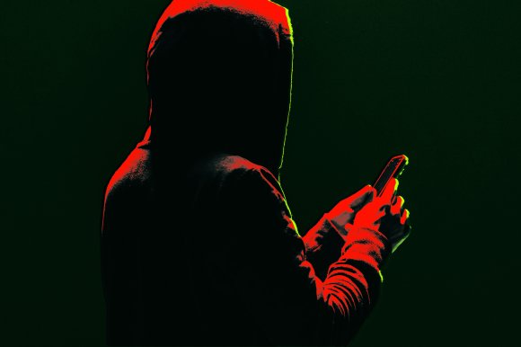 A person wearing a hooded sweatshirt is holding and looking at a smartphone. The image is dark with dramatic red and green lighting highlighting the edges of the figure and the phone. The person's face is not visible.