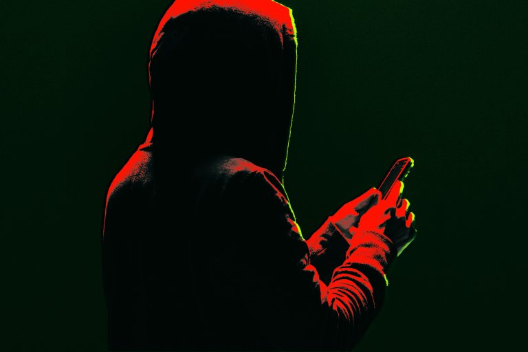 A person wearing a hooded sweatshirt is holding and looking at a smartphone. The image is dark with dramatic red and green lighting highlighting the edges of the figure and the phone. The person's face is not visible.