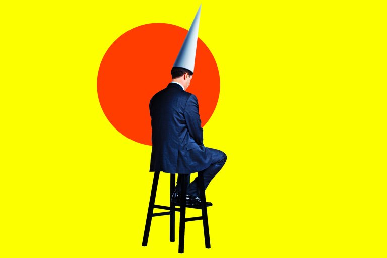 A man in a dark suit sits on a tall black stool facing away, wearing a tall white dunce cap. The background is bright yellow with a large red circle behind him.