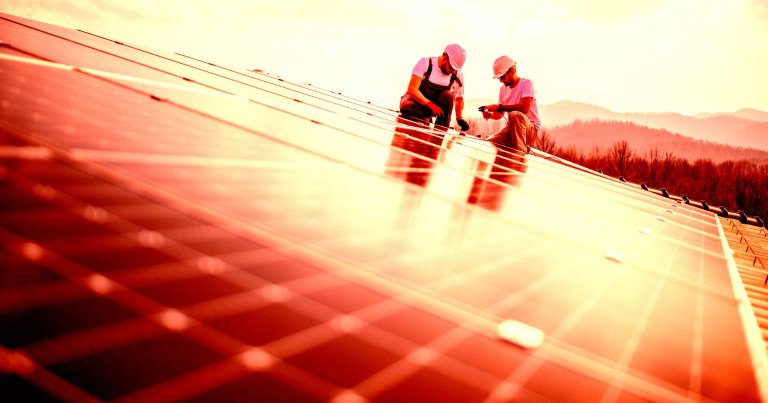 Two workers wearing white helmets and casual work clothes are installing or inspecting solar panels on a rooftop, with a backdrop of trees and distant hills under a bright sky.