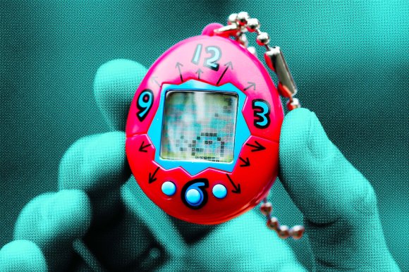 A close-up of a hand holding a red digital pet keychain toy with a small screen in the center. The toy has a clock-like design with numbers 12, 3, 6, and 9 around the screen and small arrows pointing outward. It also has three blue buttons below the screen and a silver ball chain attached at the top. The hand is wearing a teal glove.