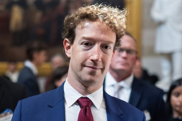 Mark Zuckerberg wearing a blue suit jacket, white dress shirt, and red tie, with curly hair, standing indoors with blurred people and artwork in the background.