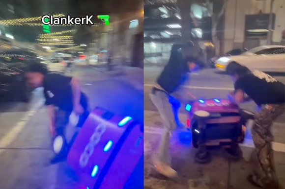 Two people are beating a tipped-over pink robot with blue lights on a city sidewalk at night. One person is wearing a black shirt and camouflage pants, while the other is dressed in a black jacket and light-colored pants. The scene is illuminated by streetlights and nearby vehicles.