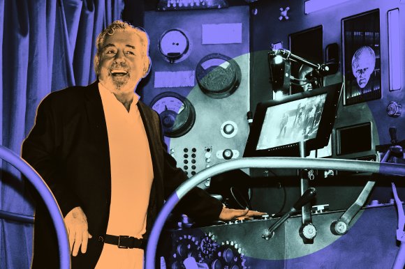 Knicks owner Jim Dolan, wearing a black jacket and white shirt, stands smiling next to a control panel with various dials, gauges, and levers.
