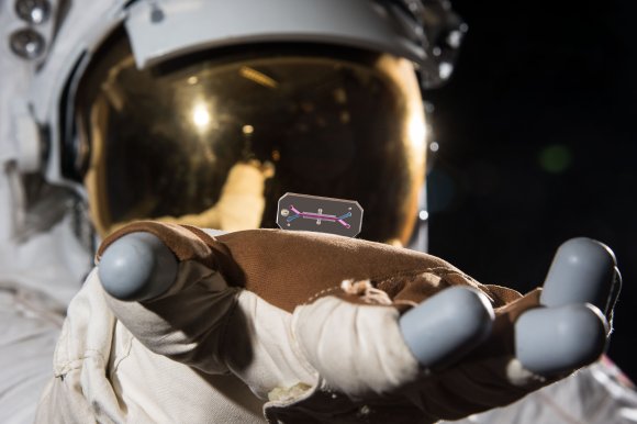An astronaut wearing a white spacesuit and gold-tinted helmet visor is holding a small, rectangular transparent chip with colored lines and dots on it, resting on the gloved hand. The background is dark, suggesting space.