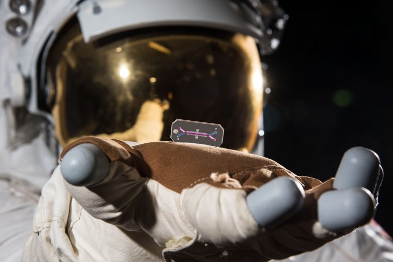 An astronaut wearing a white spacesuit and gold-tinted helmet visor is holding a small, rectangular transparent chip with colored lines and dots on it, resting on the gloved hand. The background is dark, suggesting space.
