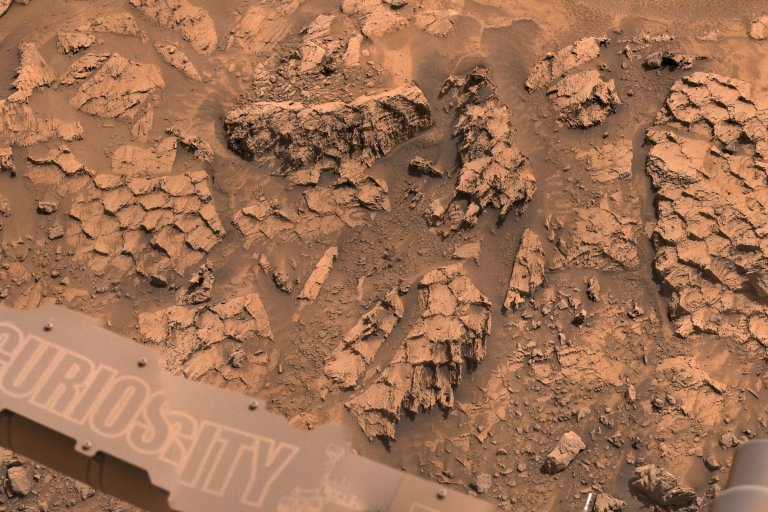 Rocky, cracked terrain with a reddish-brown hue, featuring fragmented and weathered rock formations. The lower left corner shows part of a mechanical structure with the word "CURIOSITY" partially visible, indicating the image was taken by NASA's Mars rover Curiosity.