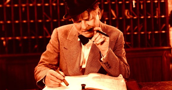A man wearing a checkered suit jacket, white shirt, black bow tie, and a black hat is sitting at a desk, holding a cigar in one hand and a pen in the other. He appears to be concentrating on writing or reviewing a large document or ledger in front of him. The background features a wooden structure with cubbyholes or mail slots. The image has a warm, reddish-orange tint.
