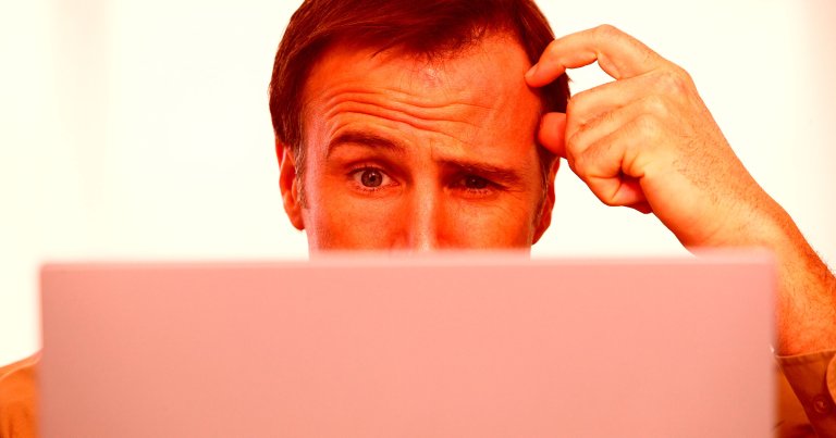 A man with short brown hair is looking at a laptop screen with a confused expression, scratching his head. The background is plain and light-colored.