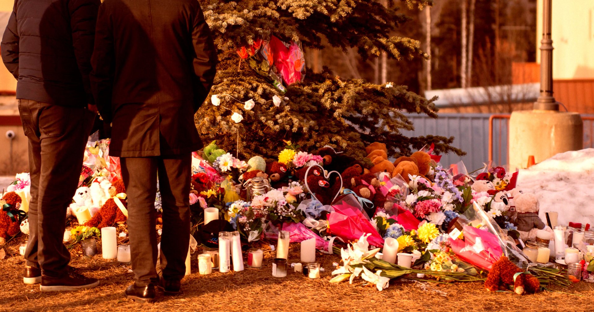 A memorial site with numerous flowers, candles, and stuffed animals placed at the base of a tree. Two people dressed in dark coats and pants stand nearby, facing the memorial. The scene is outdoors with some snow visible in the background and a warm, golden light illuminating the area.