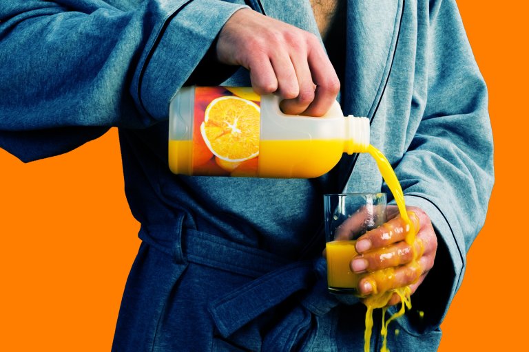 A person wearing a blue robe is pouring orange juice from a plastic bottle into a glass, but missing the glass and spilling it down their hand. The background is solid orange.