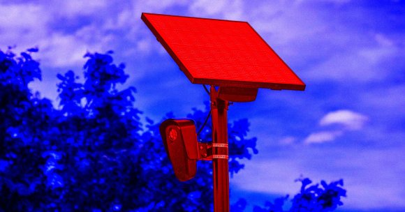 A solar panel mounted on a pole with an attached security camera, set against a background of trees and a partly cloudy sky. The solar panel and camera are highlighted in red, while the trees and sky have a blue tint.