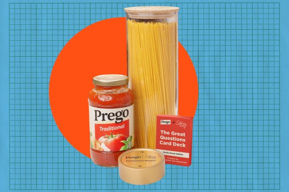 A jar of Prego Traditional Italian sauce, a tall glass container filled with uncooked spaghetti, a red box labeled "The Great Questions Card Deck Family Dinner Edition," and a beige device labeled "Prego Story Connection Keeper" are arranged together against a blue grid background with an orange circle behind them.