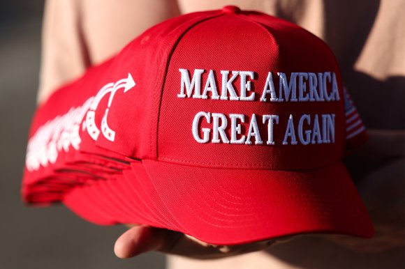 A stack of red baseball caps with white embroidered text reading "MAKE AMERICA GREAT AGAIN." The caps also feature a white arrow design on the side and an American flag patch on the back. The caps are held in a person's hand.