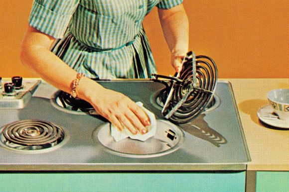 A woman wearing a green and white checkered dress is cleaning an electric stovetop. They are holding a coil burner in one hand and wiping the stovetop surface with a cloth in the other hand. The stovetop has multiple coil burners, and a cup and saucer are placed on the counter nearby. The background is a solid orange color.