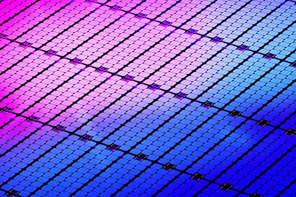Close-up of a solar panel surface showing a grid of small, rectangular photovoltaic cells. The cells display a gradient of colors from pink and purple on the left to blue on the right, with black lines separating the cells and small metallic connectors visible at intervals.