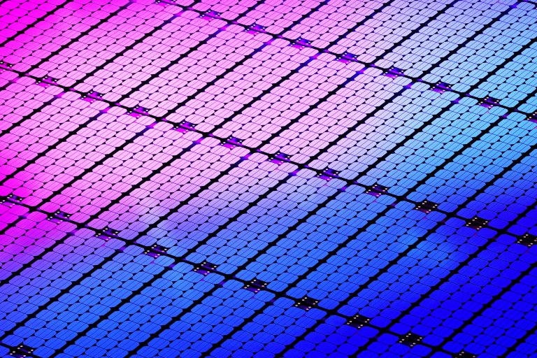 Close-up of a solar panel surface showing a grid of small, rectangular photovoltaic cells. The cells display a gradient of colors from pink and purple on the left to blue on the right, with black lines separating the cells and small metallic connectors visible at intervals.