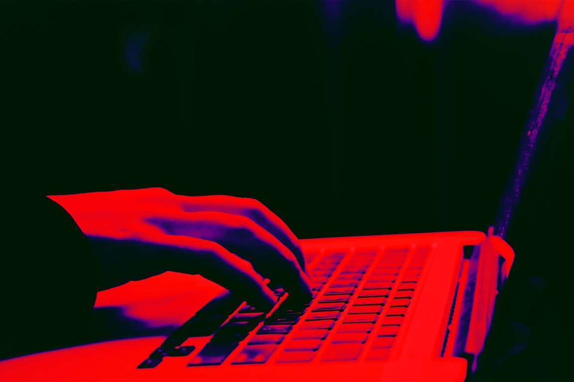 A close-up of a hand typing on a laptop keyboard, with the image rendered in a high-contrast red and dark blue color scheme. The background is dark, emphasizing the hand and keyboard.