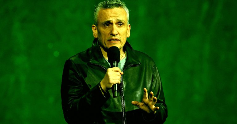 Joe Russo is shown wearing a black leather jacket is holding a microphone and speaking, with a green background behind him.