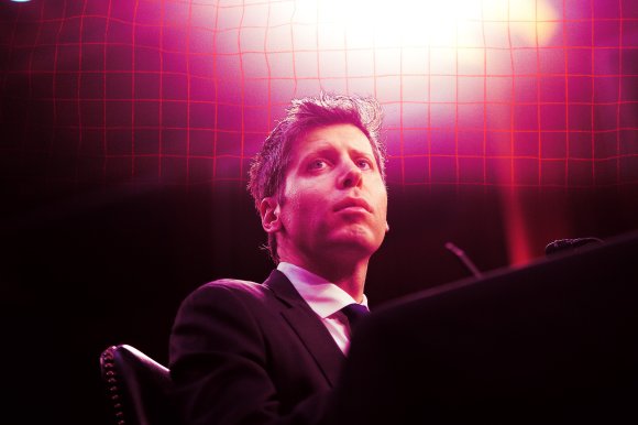 OpenAI CEO Sam Altman is seated, looking slightly upward with a serious expression. The background features a red grid pattern illuminated by pink and purple lighting, creating a dramatic and intense atmosphere.