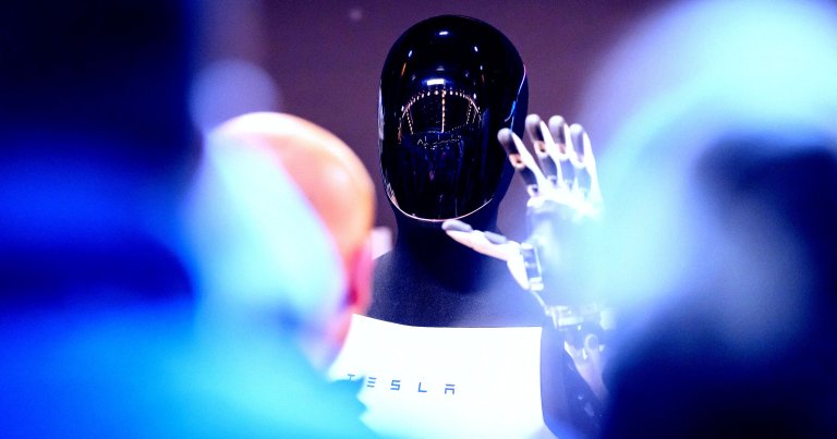A black humanoid robot with a glossy, reflective faceplate and a mechanical hand raised in a waving gesture. The robot is wearing a white garment with the word "TESLA" printed on it. The image has a blue and purple lighting effect, with blurred figures in the foreground.
