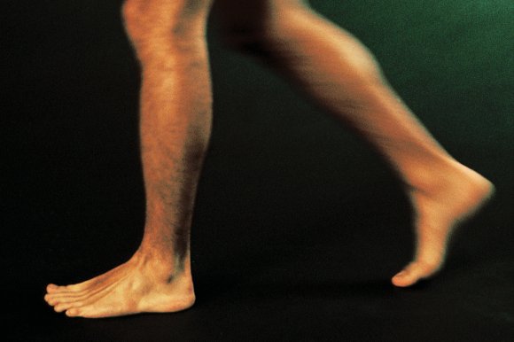 Two bare legs and feet of a person walking or running, with one foot flat on the ground and the other foot raised on the toes, set against a dark background.