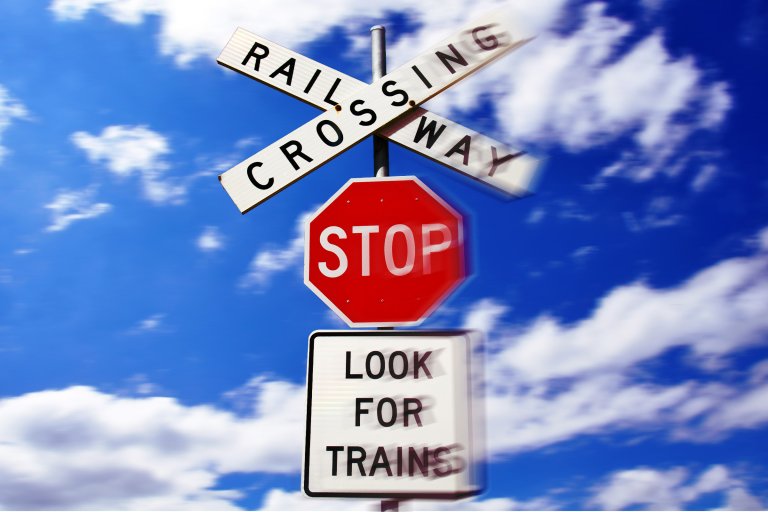 Railroad crossing sign with "RAILROAD CROSSING" in black letters on white crossbuck boards, a red octagonal stop sign below it, and a white rectangular sign underneath that reads "LOOK FOR TRAINS," all set against a blue sky with scattered white clouds. The signs appear slightly blurred, suggesting motion or vibration.