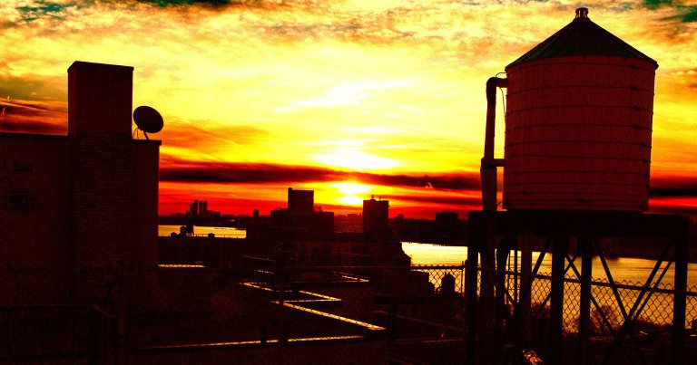 Sunset over a city skyline with silhouetted buildings, a water tower on the right, and a satellite dish on a rooftop chimney on the left. The sky is filled with vibrant yellow, orange, and red hues reflecting on the water below.