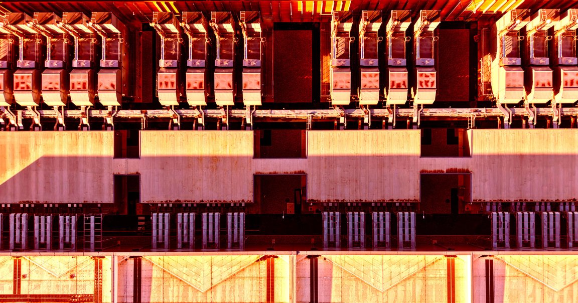Aerial view of a large industrial rooftop with multiple rows of HVAC units and ventilation systems. The image is bathed in warm, orange sunlight, casting long shadows and highlighting the geometric patterns of the equipment and rooftop structures. The rooftop appears to be part of a commercial or industrial building.